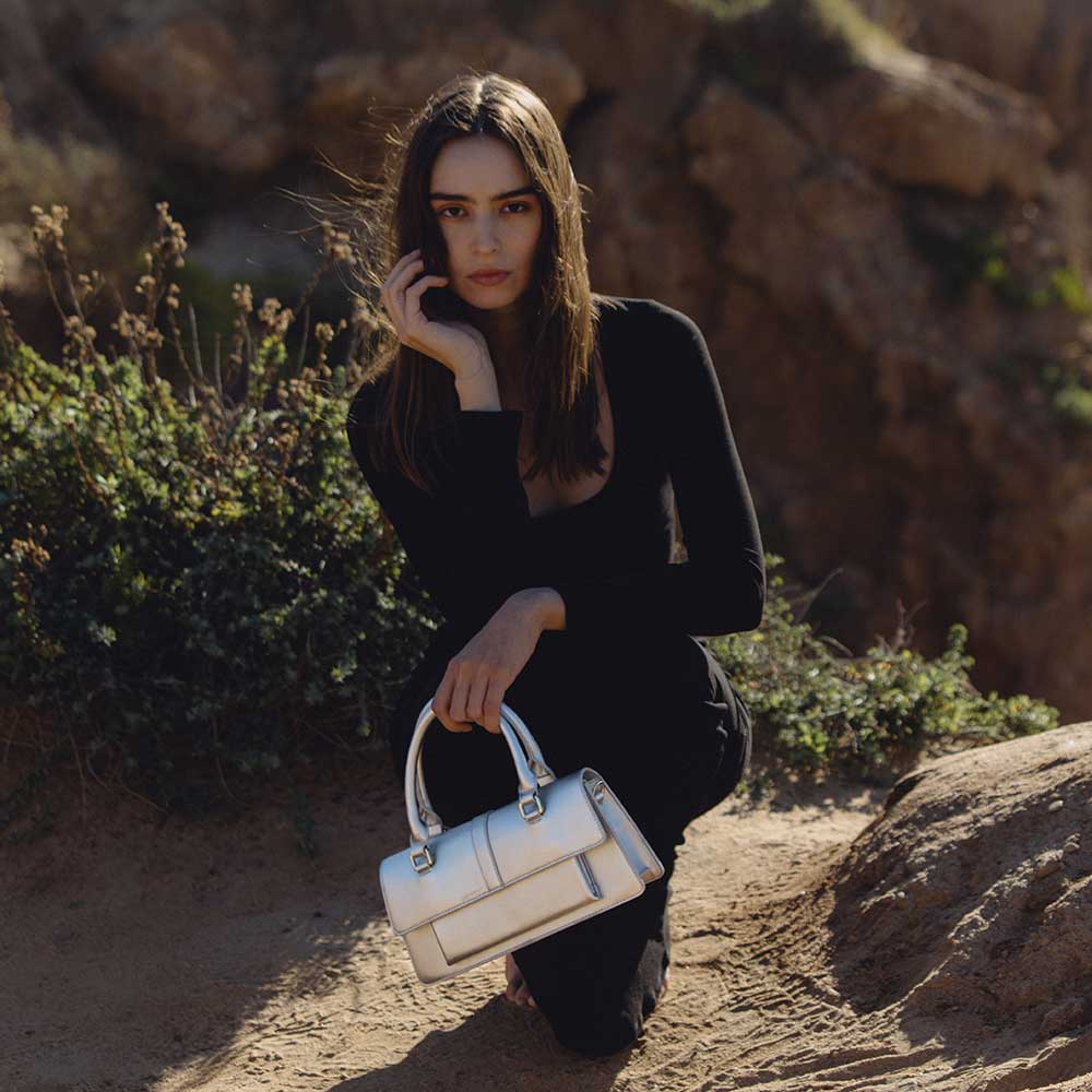 A model wearing a small silver vegan leather top handle bag while sitting outside.