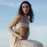 A model wearing a small tan woven vegan leather crossbody bag with a drawstring closure while sitting outside.